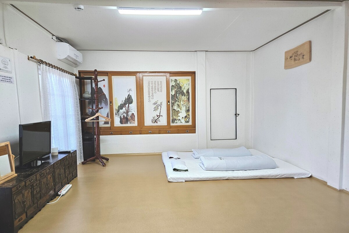 Une pièce avec un lit, un téléviseur posé sur une commode, un porte-manteau et une porte.