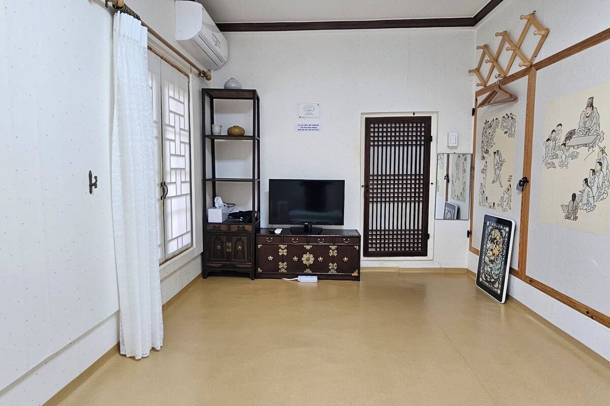 A room with a TV on a wooden cabinet, a white door with a curtain, a wall-mounted air conditioner, and a traditional door with a grid pattern.