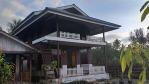 A two-story wooden house with a balcony, surrounded by vegetation.