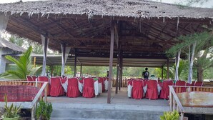 An outdoor covered seating area with tables set for dining, a thatched roof, and a view of the surrounding greenery.