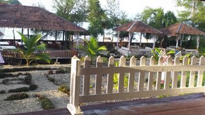 A wooden deck with a white picket fence, surrounded by small plants and a thatched-roof structure.