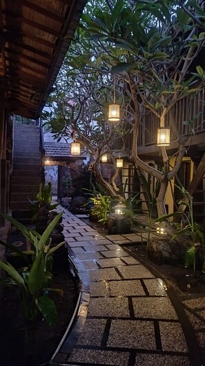 A pathway leading to a wooden structure with hanging lights, surrounded by greenery and stone elements.