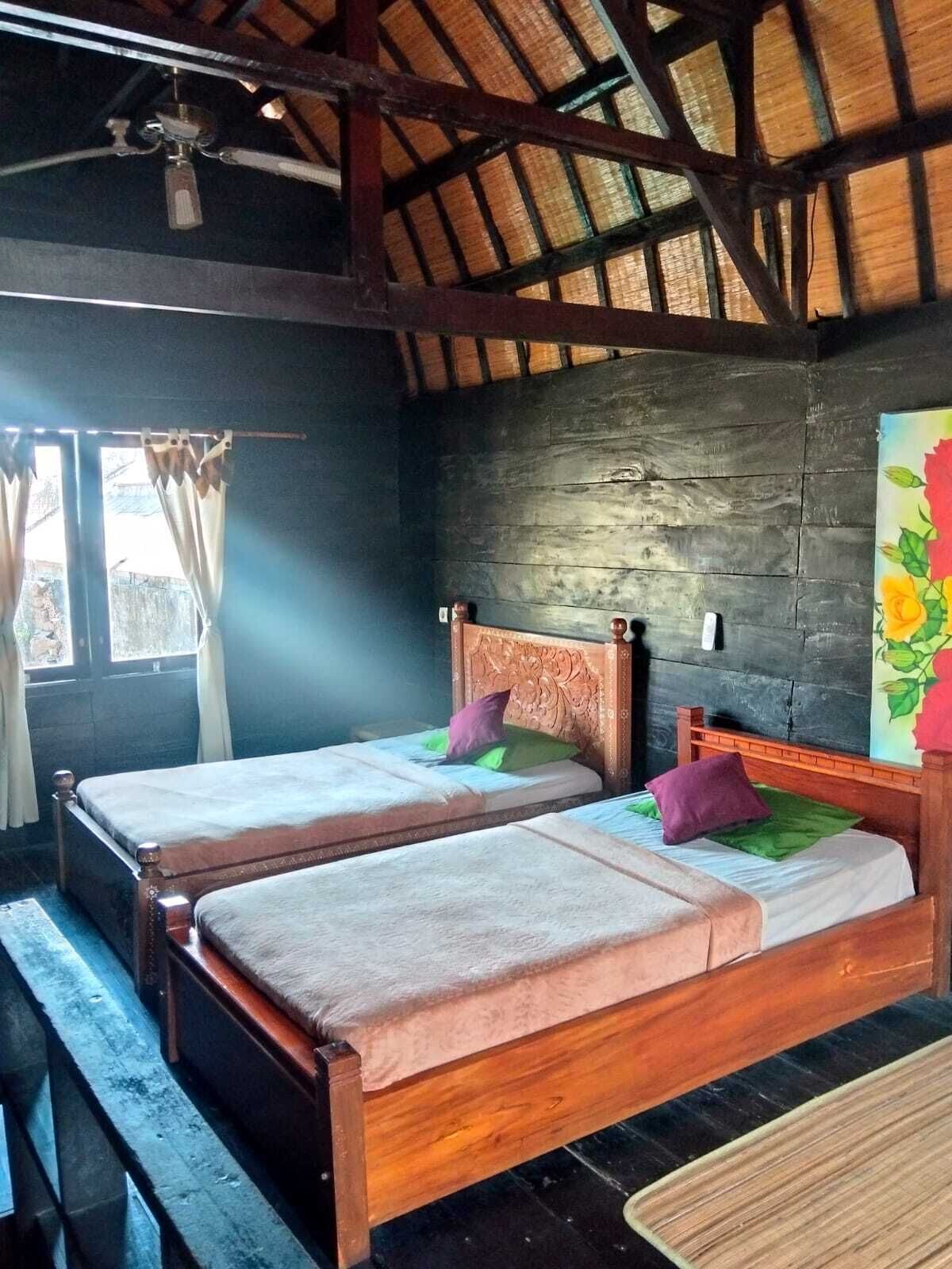 Une pièce avec deux lits en bois, un ventilateur de plafond, une fenêtre avec des rideaux et un tableau au mur.