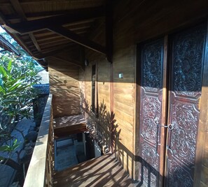 A wooden building with a balcony, a door with intricate carvings, and a small garden area with plants and a stone pathway. - Bale Sasak (Gili Trawangan)