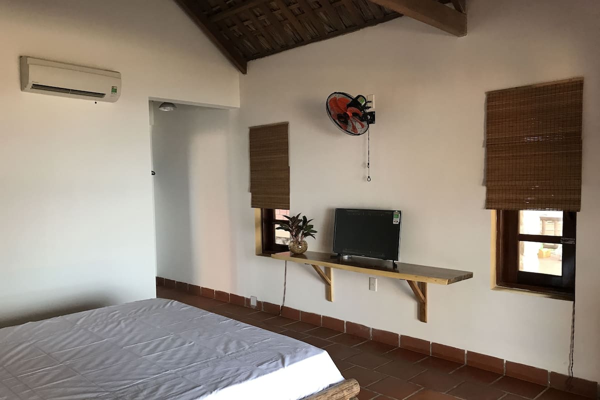 A bedroom with a bed, a TV on a shelf, a window with a blind, and an air conditioning unit.
