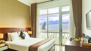 A hotel room with a large bed, a desk, a chair, and a view of the sea and mountains through the windows. - Navy Hotel Danang (Da Nang)