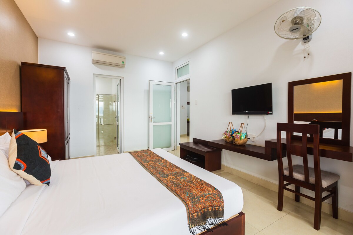 A hotel room with a bed, a desk, a chair, a TV, and a fan.