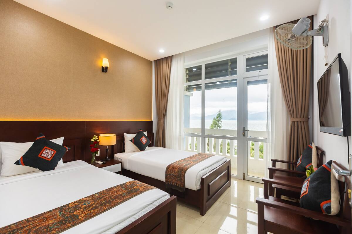 A hotel room with two beds, a TV, a balcony with curtains, and a view of the outdoors.
