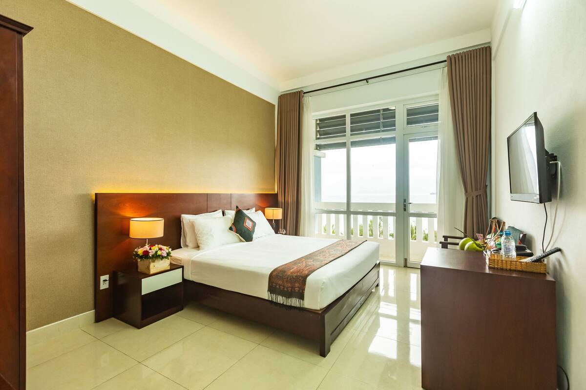 A hotel room with a large bed, a nightstand with a lamp, a desk with a TV, and a balcony with curtains.