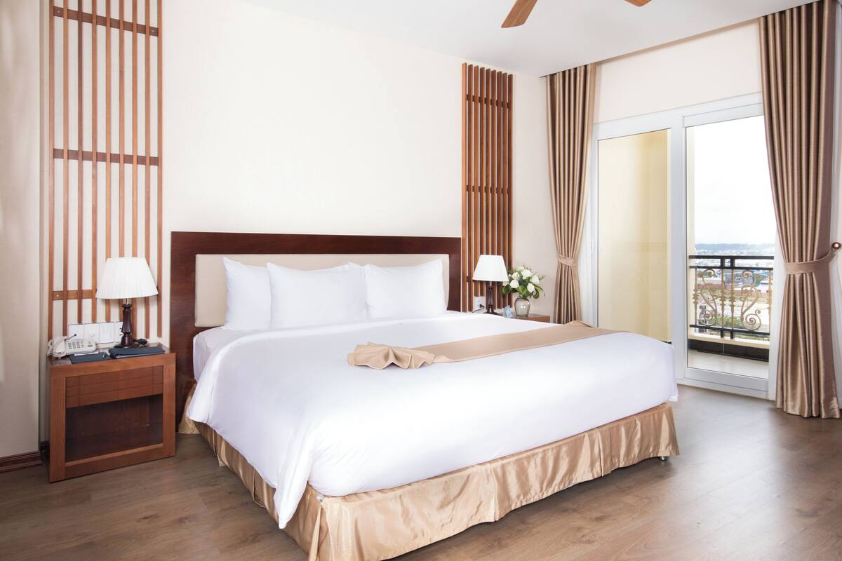 Habitación de hotel con una cama grande, cabecera de madera, mesitas de noche con lámparas y vistas al exterior a través de un ventanal corredero.