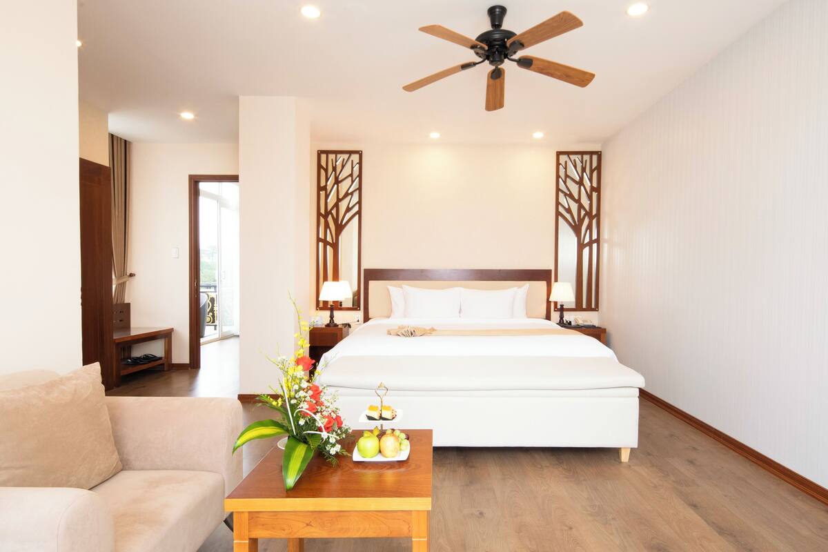 Habitación de hotel con cama, mesita de noche de madera, mesa con un florero y fruta, y ventilador de techo.