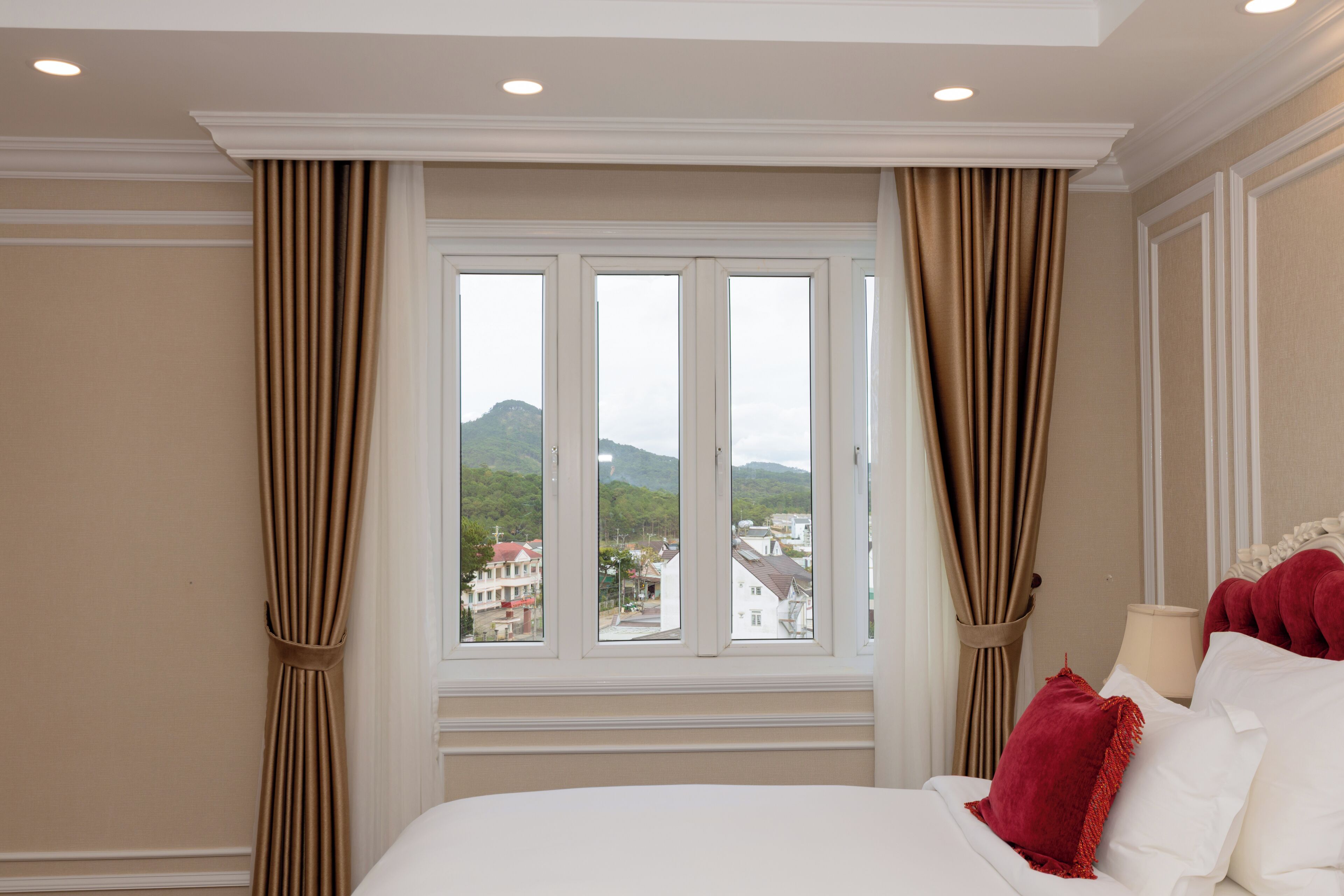 Una habitación de hotel con un ventanal grande, una cama con sábanas blancas, almohadas rojas y cortinas color beige.