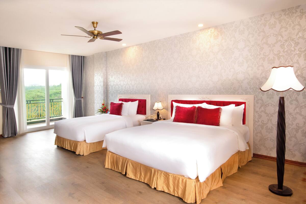 Habitación de hotel con dos camas, ventilador de techo, mesita de noche con lámpara y vista a zonas verdes.
