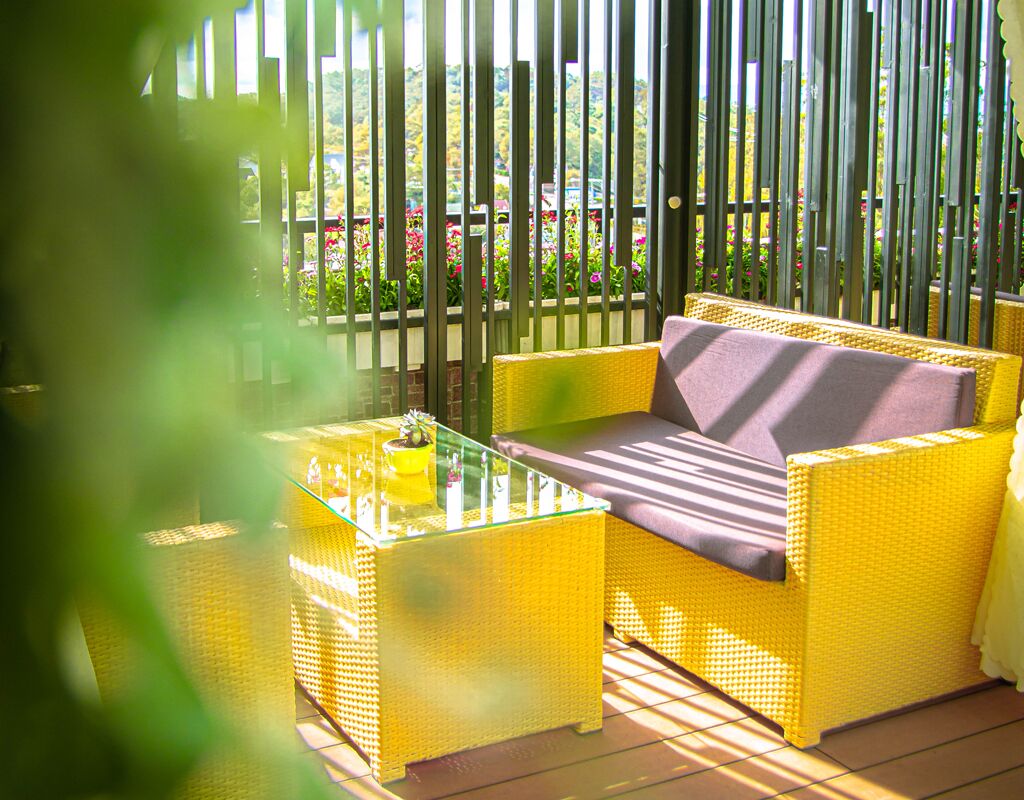 A yellow wicker sofa with a cushion, a glass coffee table, and a view of a garden through vertical blinds.