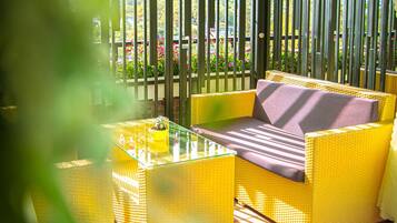 A yellow wicker sofa with a cushion, a glass coffee table, and a view of a garden through vertical blinds.