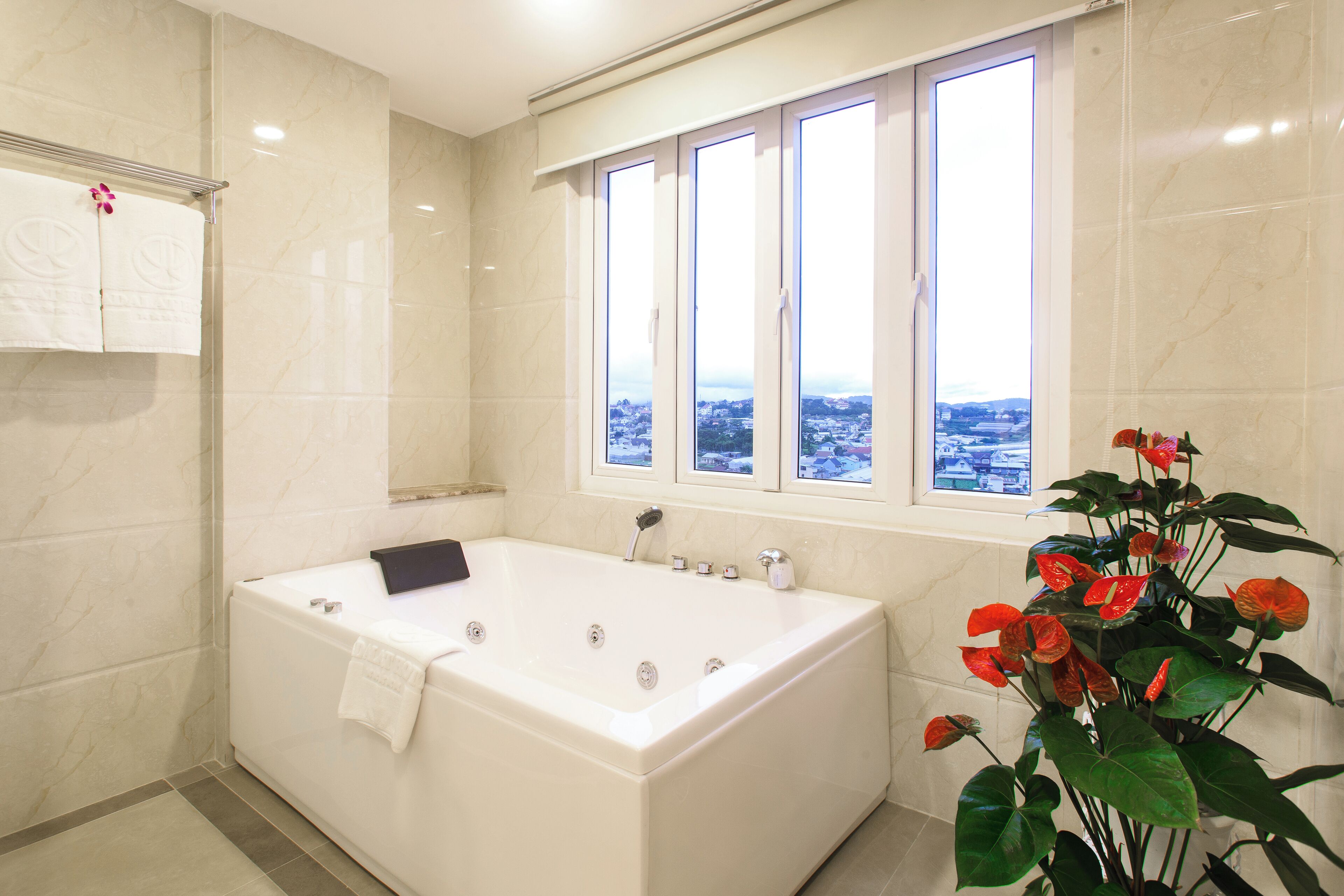 A modern bathroom with a large bathtub, a window with a city view, and a potted plant with red flowers.