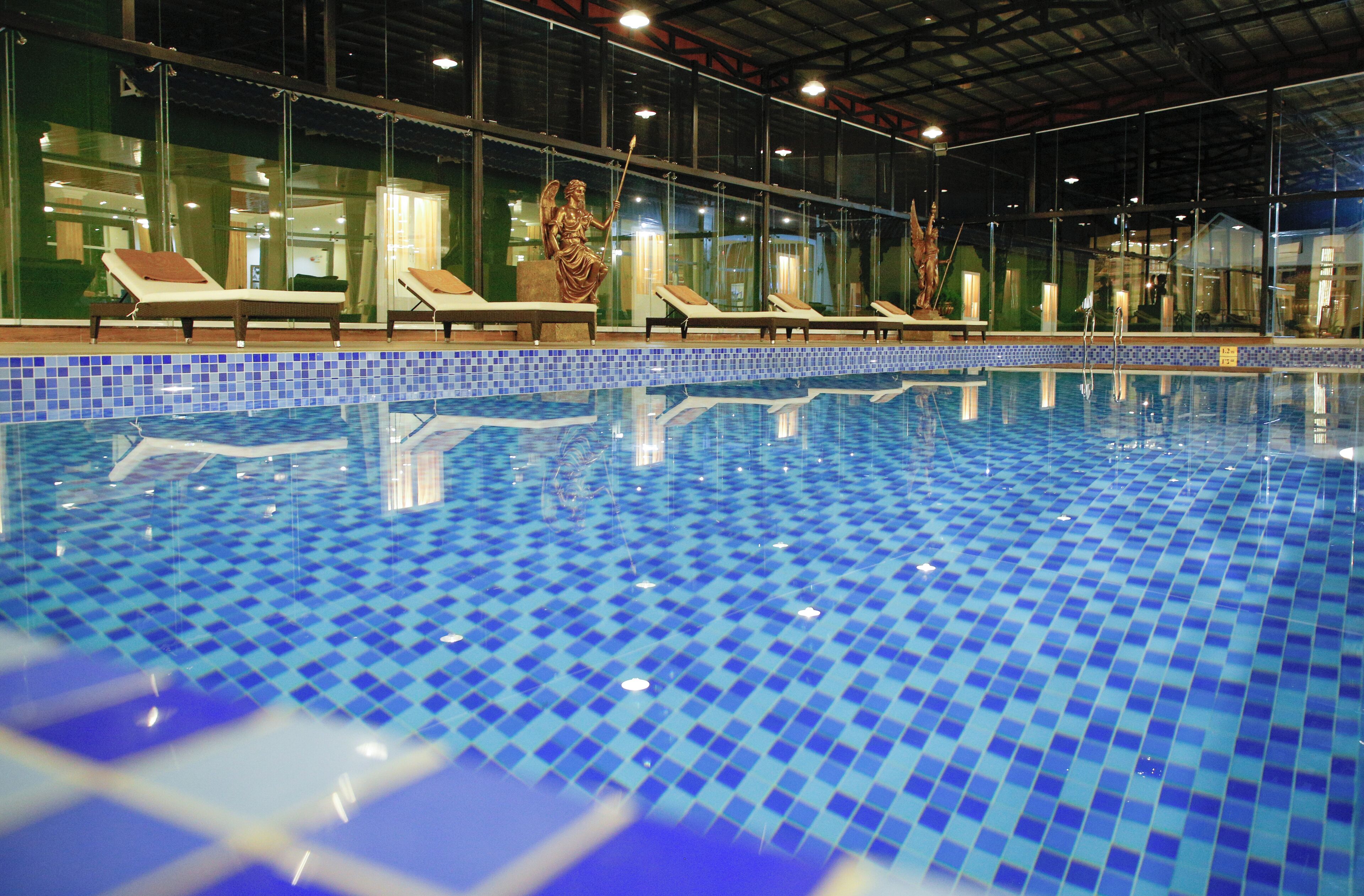 Una piscina interior con mosaicos azules, sillones de descanso y una estatua de un ángel.