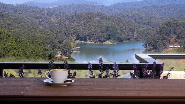 A view from a balcony with a cup of coffee and purple flowers, overlooking a lake and forested mountains. - Ladalat Hotel (Lam Vien - Da Lat)