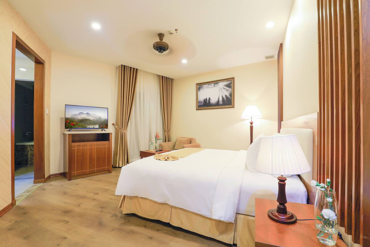 Habitación de hotel con una cama, un escritorio con lámpara, un televisor y un florero con flores.