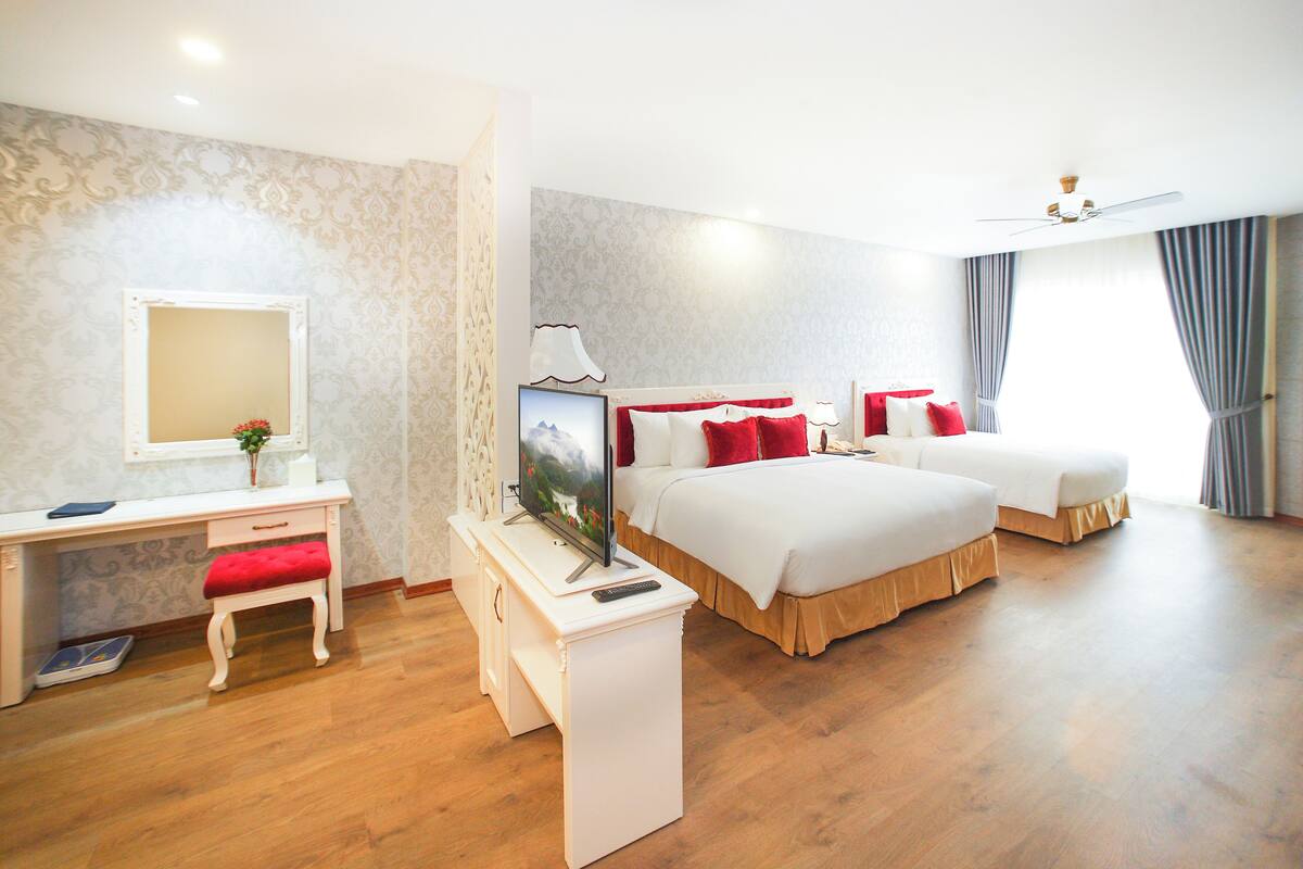 Una habitación de hotel con escritorio, dos camas, televisor y un ventanal grande con cortinas.