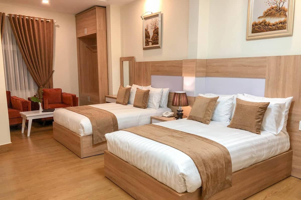 A hotel room with two beds, a wooden headboard, a desk with a lamp, a chair, and a large window with curtains.