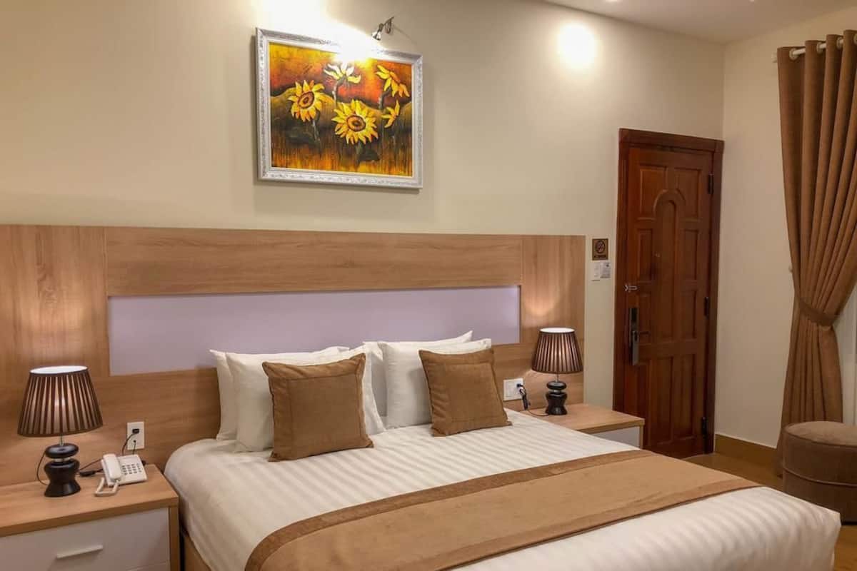 A hotel room with a large bed, two bedside tables with lamps, a wooden headboard, a painting on the wall, a door, and a curtain.