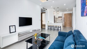 Ein modernes Wohnzimmer mit Flachbildfernseher, Couchtisch mit Vase, blauem Sofa, Deckenventilator, Einbauleuchten und einer Küchenzeile im Hintergrund.