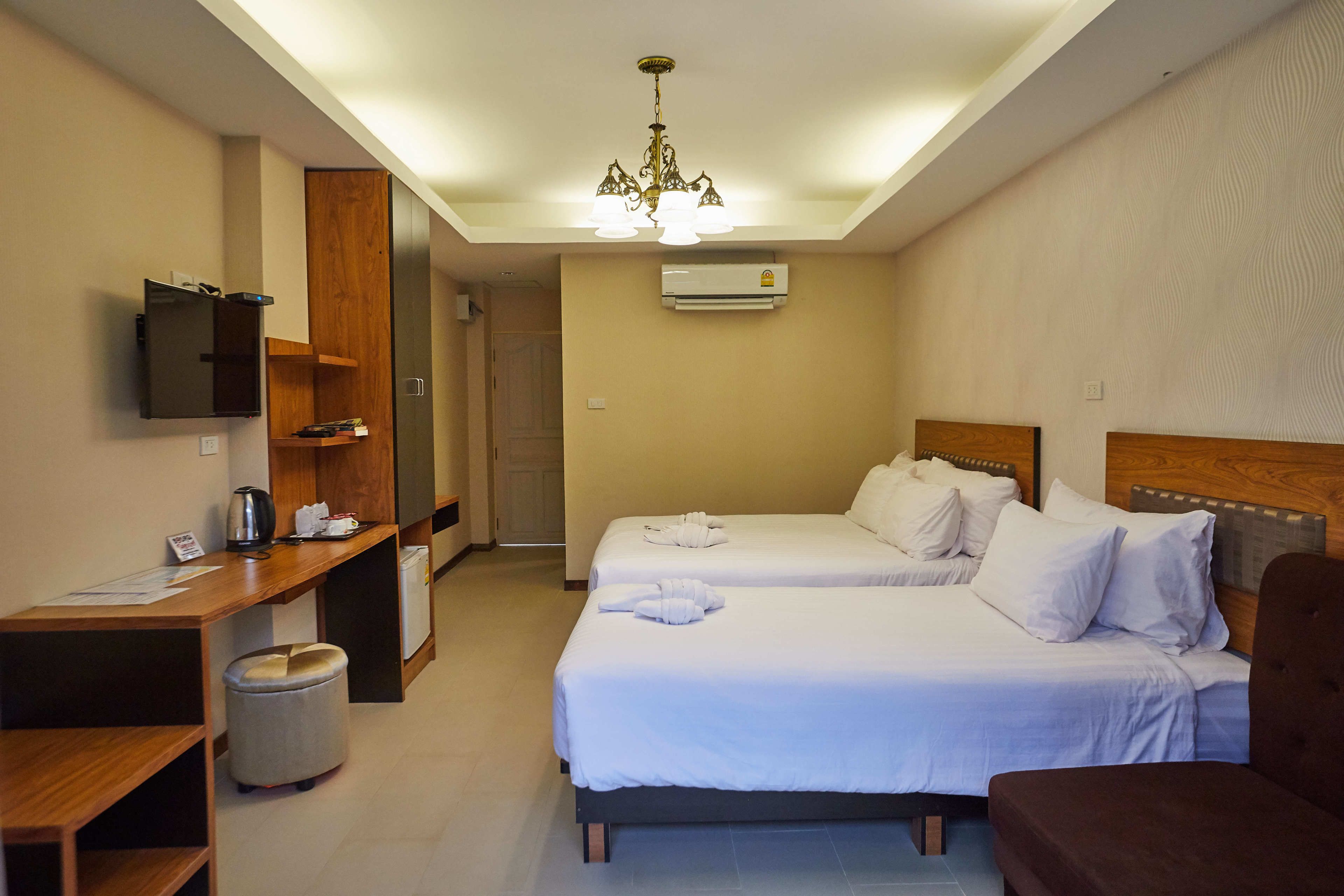 A hotel room with two beds, a desk, a chair, a trash bin, a TV, and a chandelier.