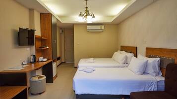 A hotel room with two beds, a desk, a chair, a trash bin, a TV, and a chandelier.