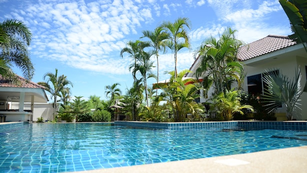 A swimming pool with tiled edges, surrounded by palm trees and a building with a tiled roof. - Bangsaray Villa (Sattahip)