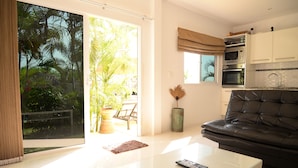 A modern living room with a glass coffee table, a black leather sofa, and a sliding glass door leading to a balcony with a view of palm trees. - Bangsaray Villa (Sattahip)