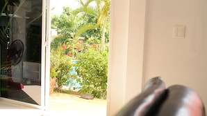 A room with a large window, a fan, and a view of a garden with various plants and a small pond. - Bangsaray Villa (Sattahip)