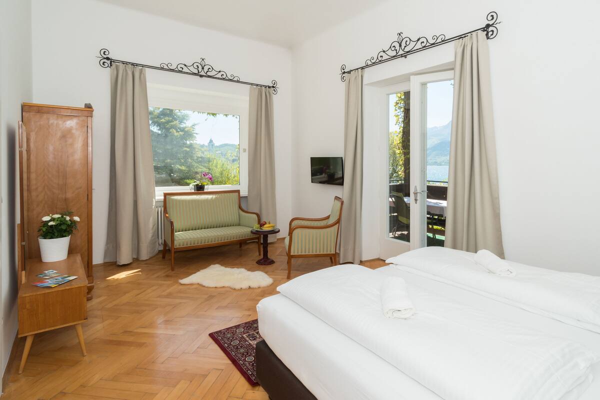 Ein Schlafzimmer mit einem großen Bett, einem Holzschrank, einem kleinen Tisch mit einer Pflanze, zwei Stühlen und Blick ins Freie durch ein Fenster.