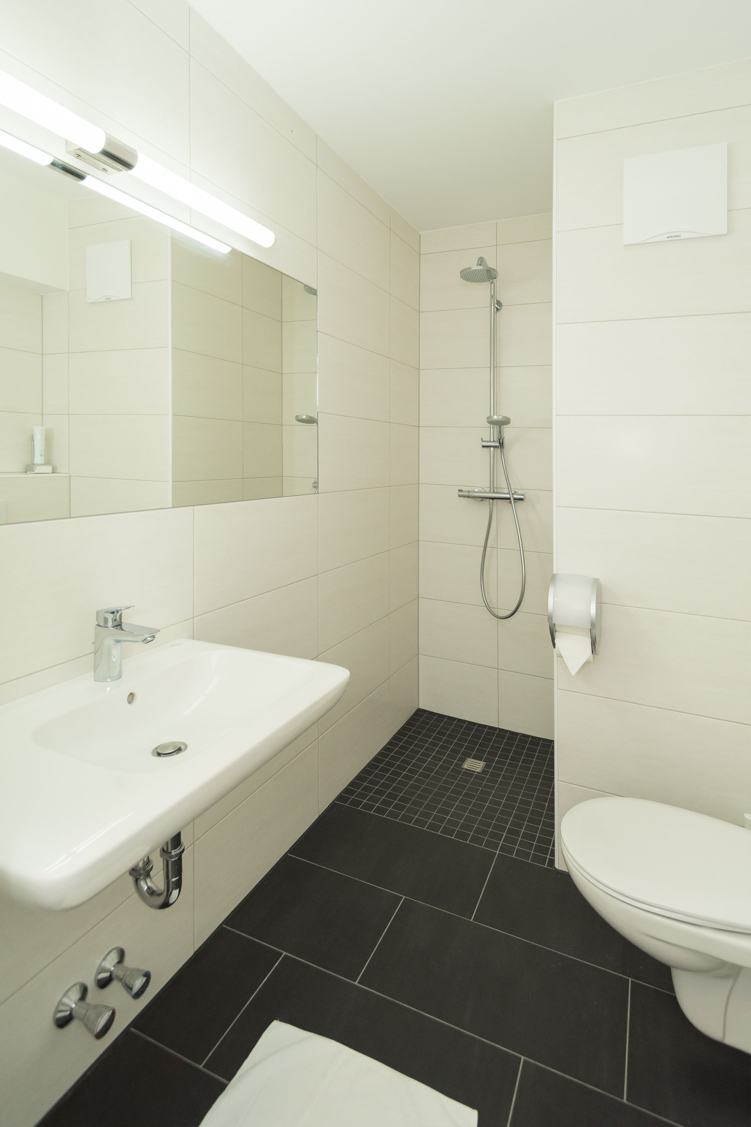Un bany modern amb un lavabo, un mirall i un vàter de color blanc, una zona de dutxa i un paviment de rajoles fosques.