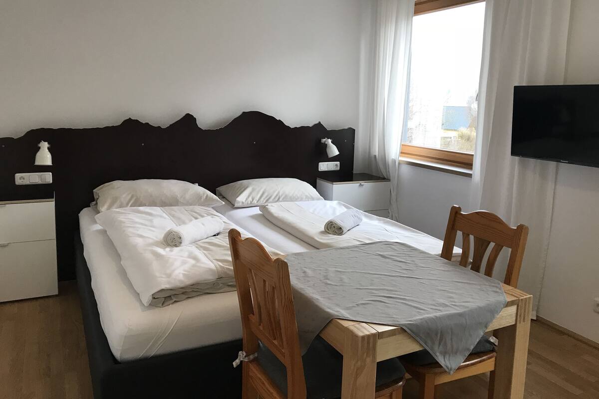 Ein ordentlich bezogenes Bett mit weißen Leinen, ein Nachttisch mit Lampe, ein an der Wand befestigter Fernseher und ein Fenster mit Vorhängen.