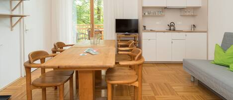 Una habitación moderna y bien iluminada con una mesa de comedor y sillas de madera, un sofá gris, una cocina con armarios blancos y un amplio ventanal con vistas a zonas verdes.
