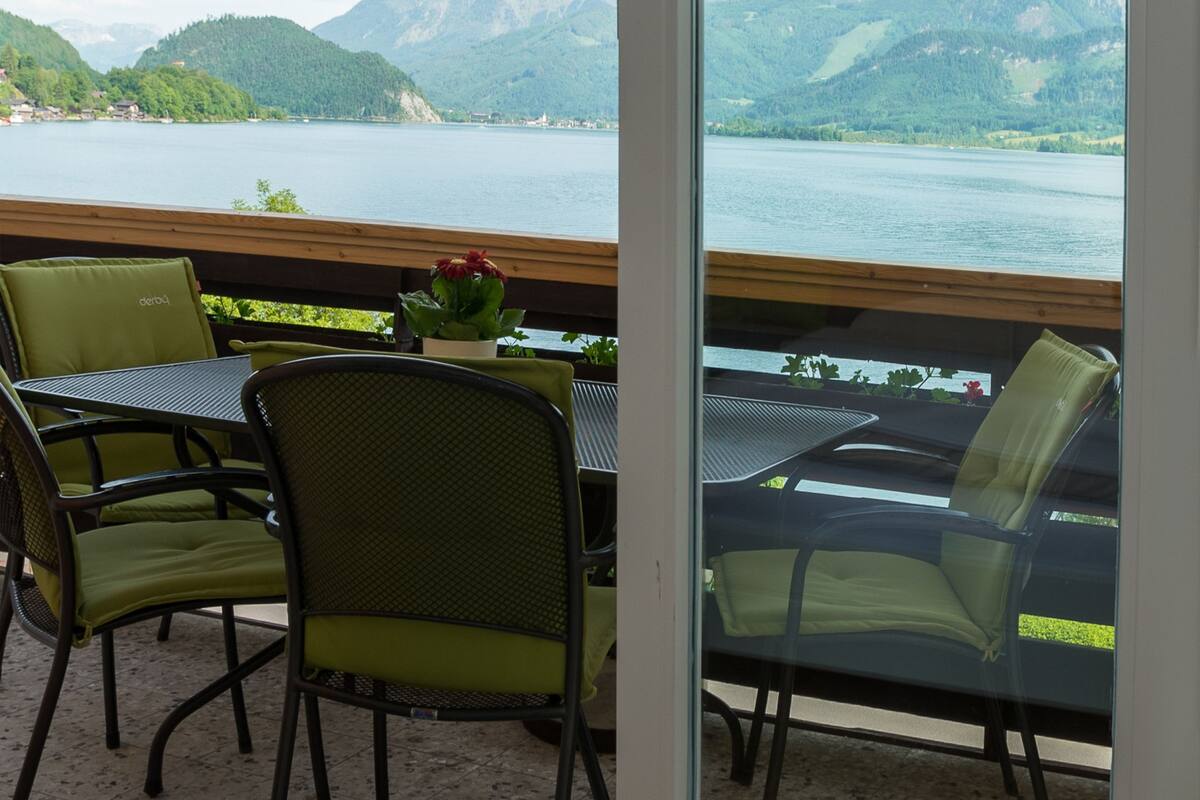 Ein Balkon mit Tisch und Stühlen, der auf einen See und Berge blickt.