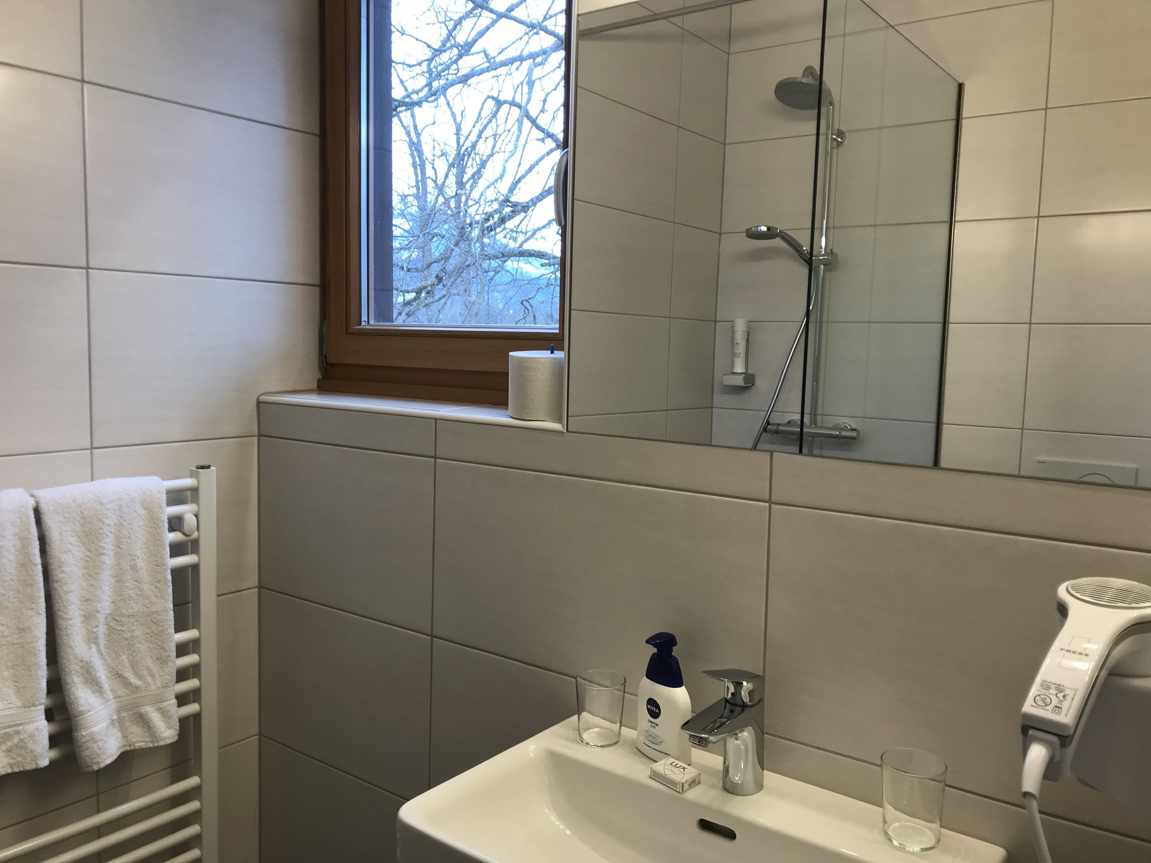 Een badkamer met een wastafel, spiegel en douche. Er is een raam boven de wastafel, en handdoeken hangen aan een rek.