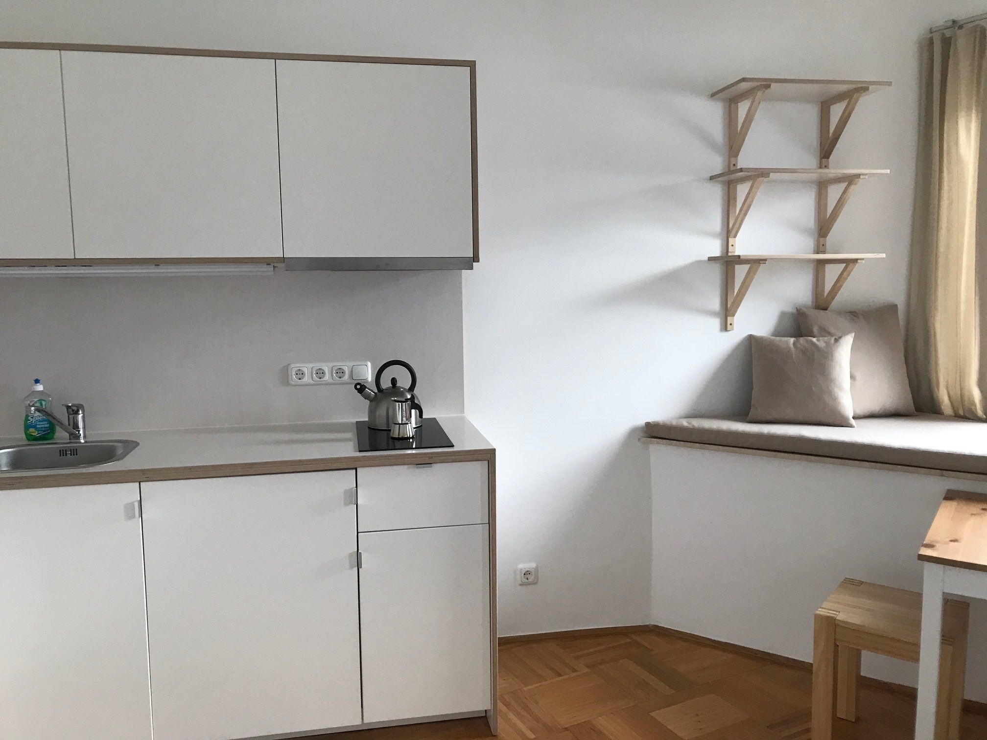 A compact kitchen with white cabinets, a kettle, and a wooden shelf unit.