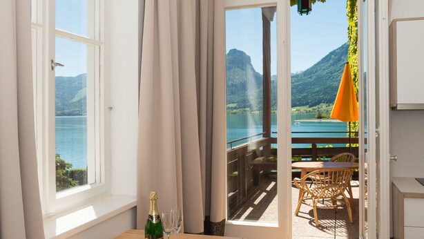 A room with a wooden table, a bottle of champagne, and two glasses. There is a view of a balcony with outdoor furniture and a mountain landscape. - Seehaus Familie Leifer (Sankt Wolfgang im Salzkammergut)