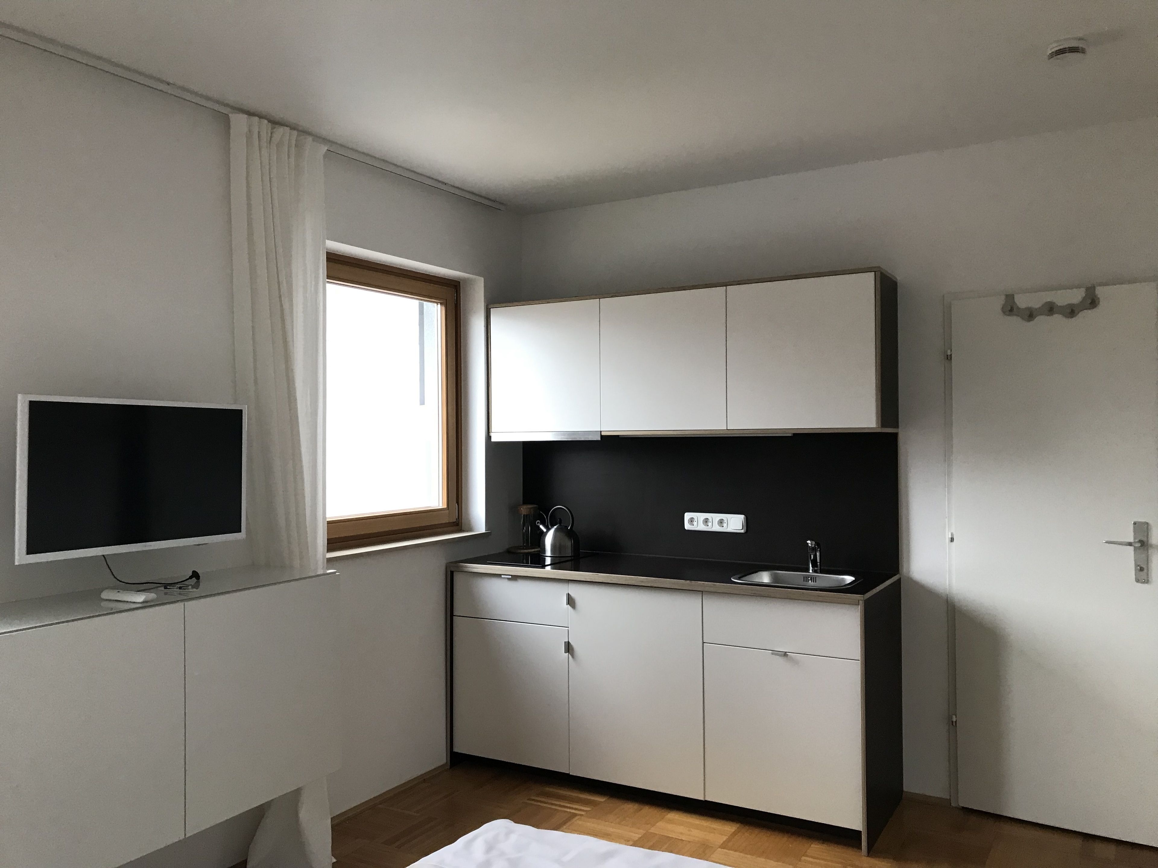 Een compacte, moderne keuken met witte kasten, een zwart aanrecht, een raam met gordijnen, een ingebouwd spoelbak en een flatscreen televisie.