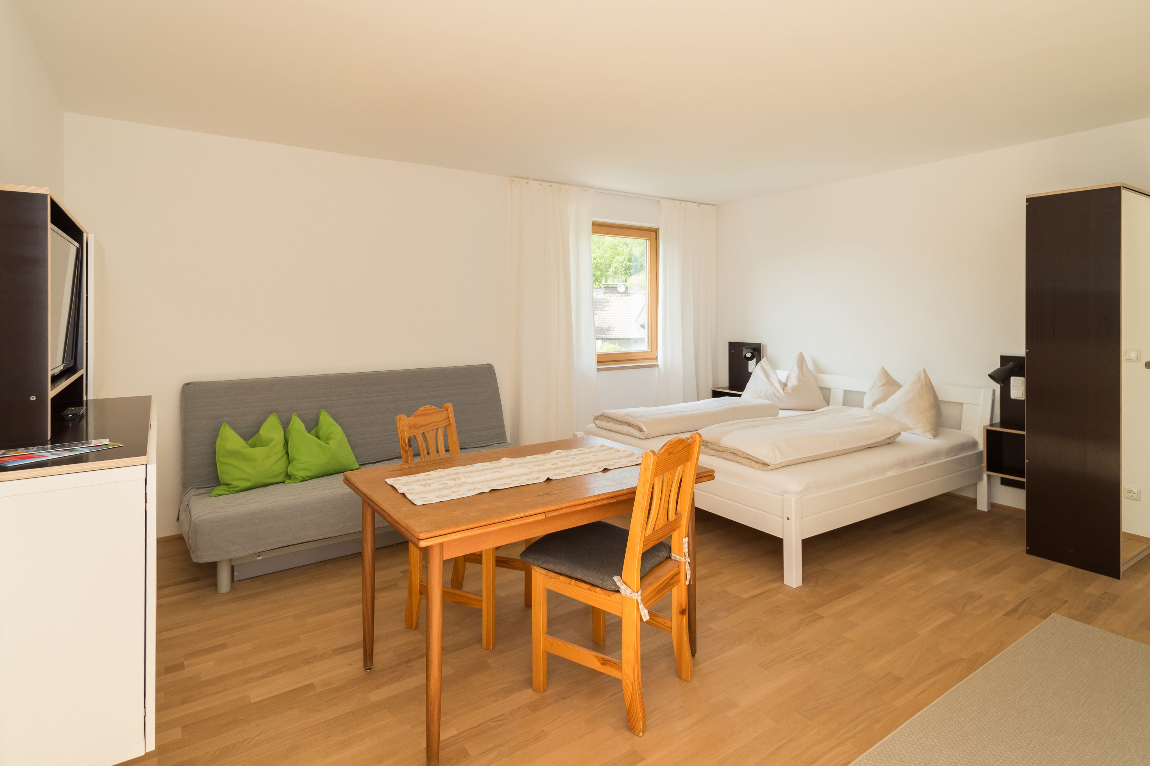 Een compacte kamer met een houten eettafel, een grijze bank met groene kussens, een wit bed en een donkere garderobe.