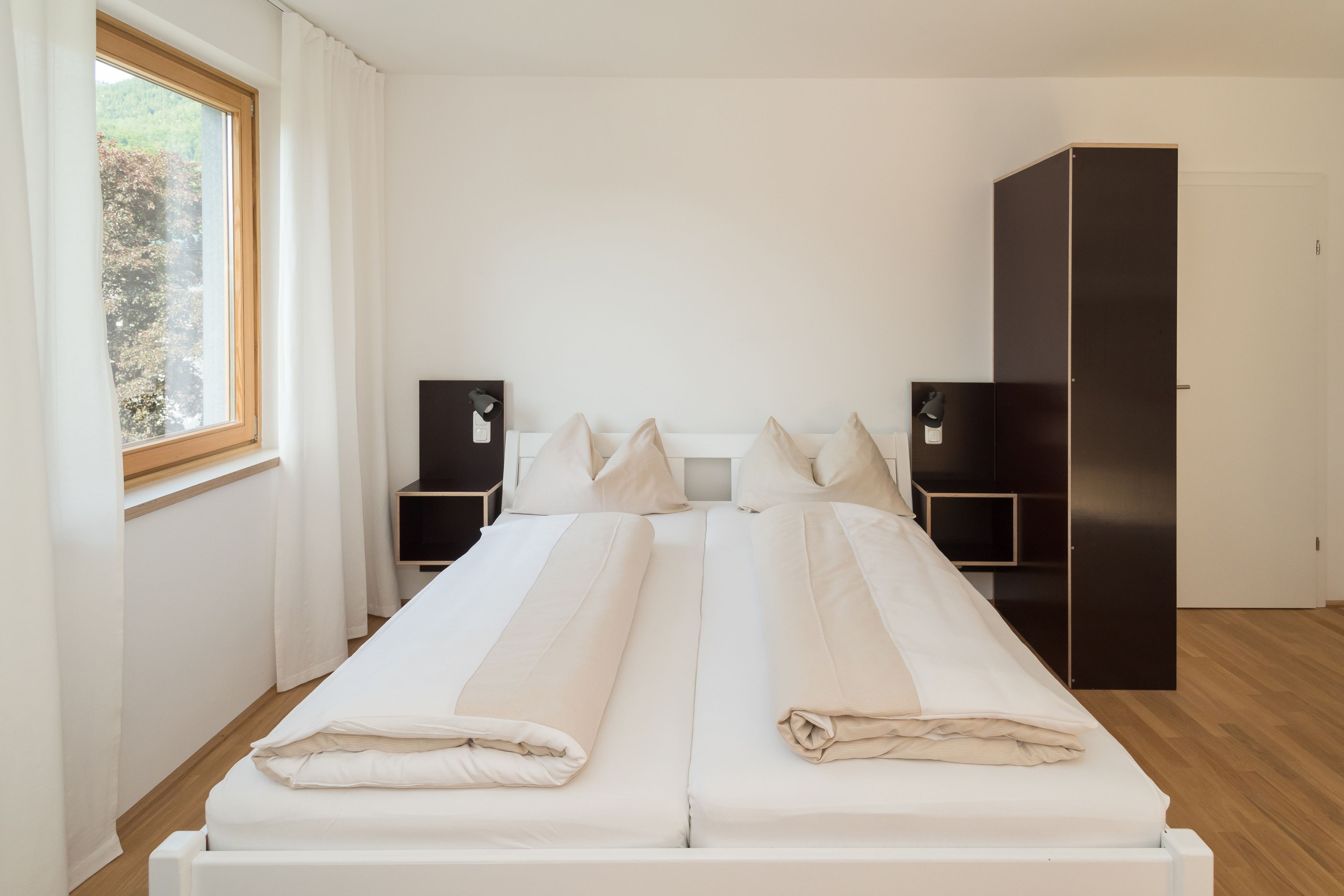 A double bed with white linens, a bedside table with a lamp, a wardrobe, and a window with curtains.