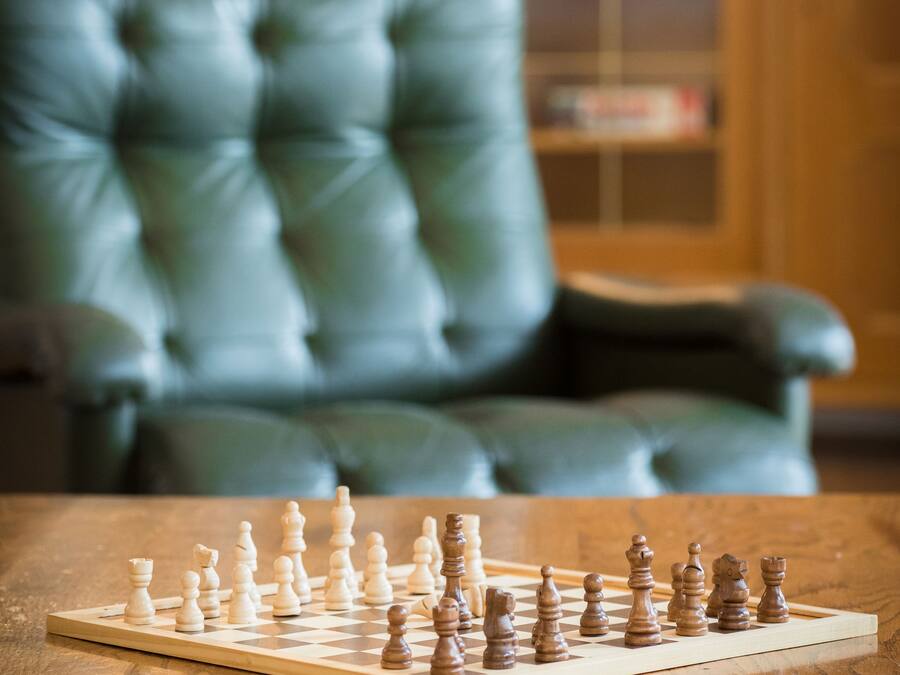 Um tabuleiro de xadrez sobre uma mesa de madeira, com uma cadeira de couro verde ao fundo.