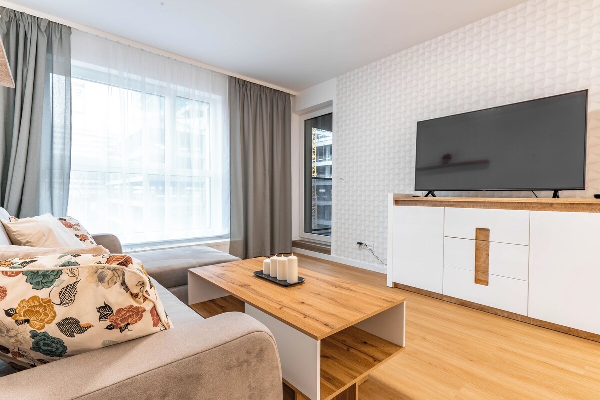 A modern living room with a sofa, a wooden coffee table, a TV, and a window with curtains.
