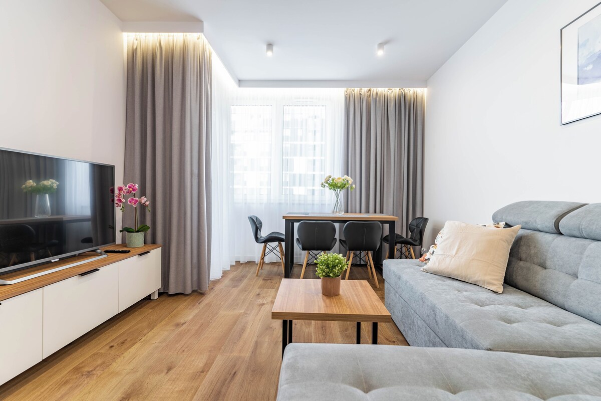 A modern living room with a flat-screen TV, a grey sofa, a wooden coffee table, and a dining area with a table and chairs.