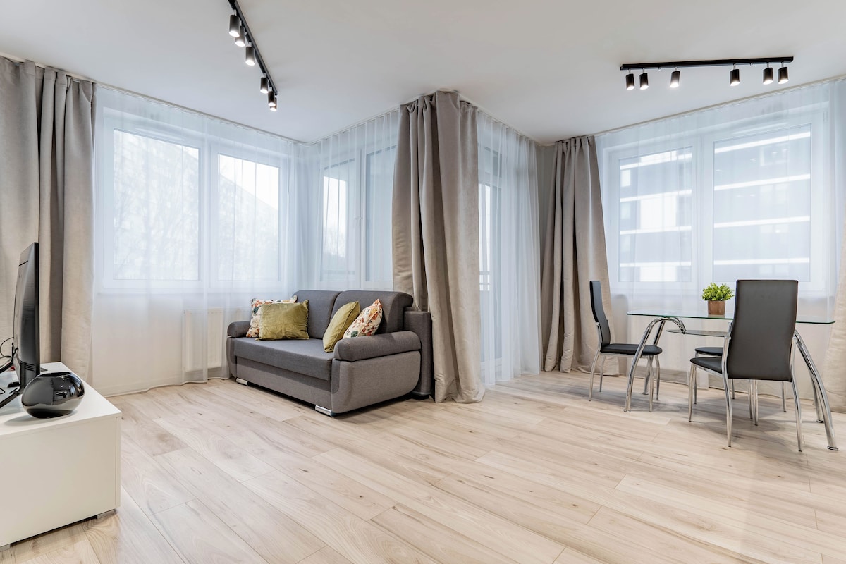 A modern living room with a grey sofa, a glass coffee table, and a dining area with two chairs.
