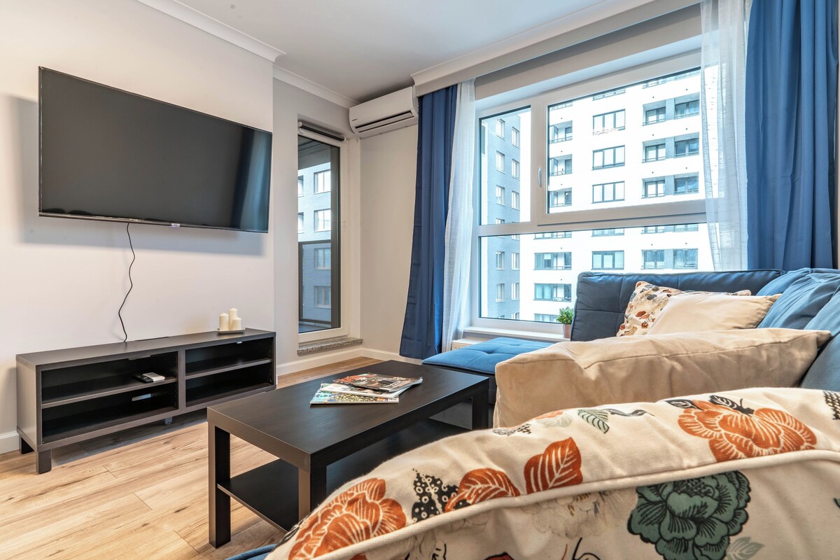 A modern living room with a flat-screen TV, a dark wooden coffee table, a sofa with patterned cushions, and a large window with curtains.