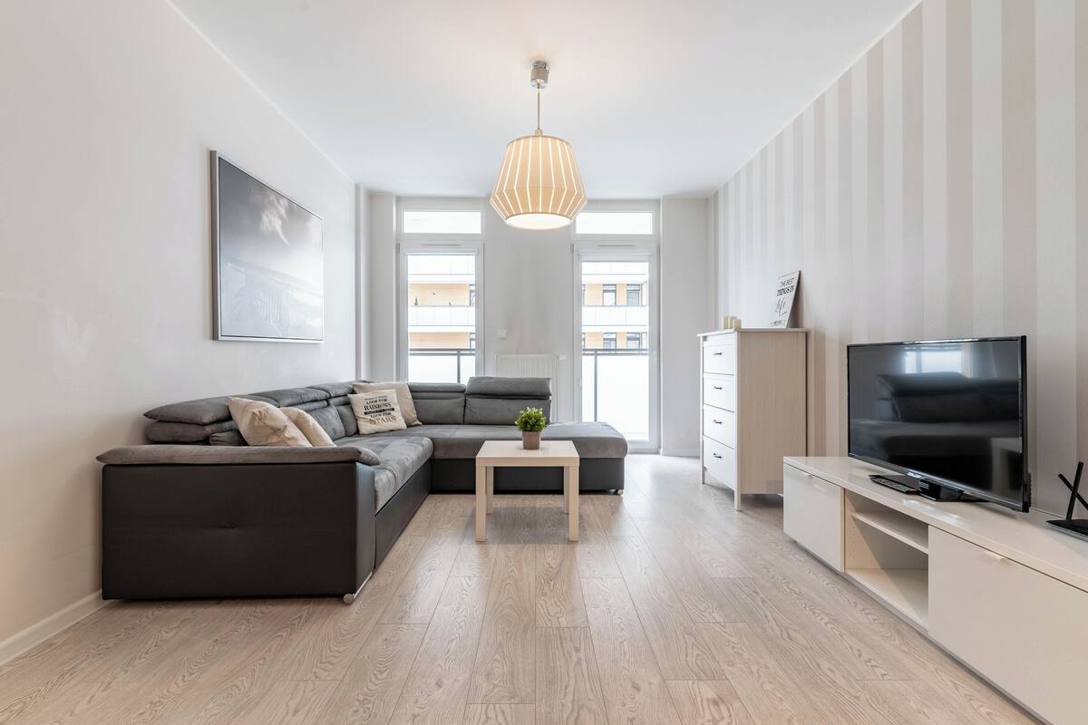 Una sala de estar moderna con un sofá modular, una mesita con una planta, un televisor sobre un mueble bajo y un ventanal grande.