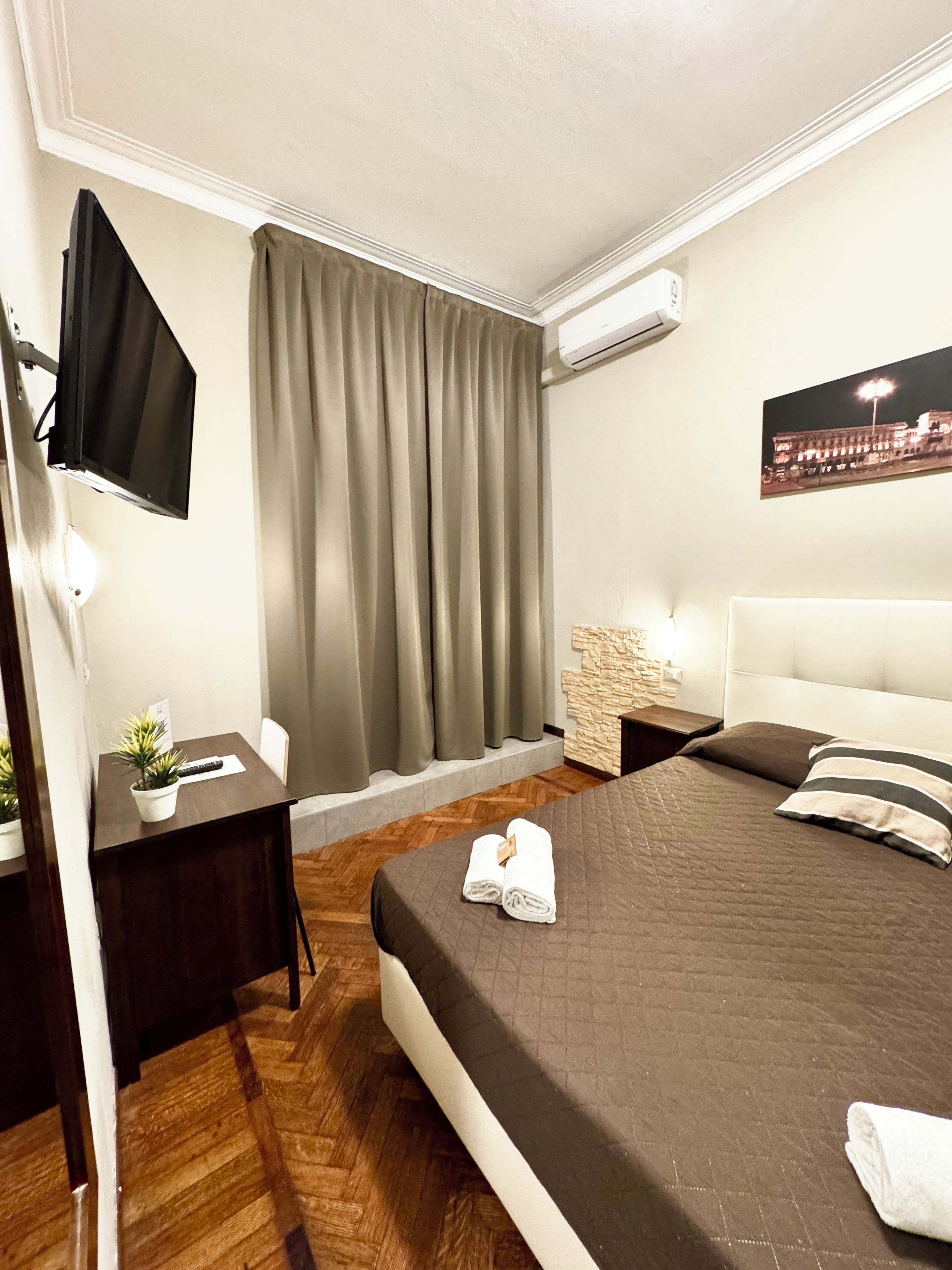 Ein Hotelzimmer mit einem Bett, einem an der Wand montierten Fernseher, einem kleinen Tisch mit einer Pflanze, einem Nachttisch und einem Stadtbild.